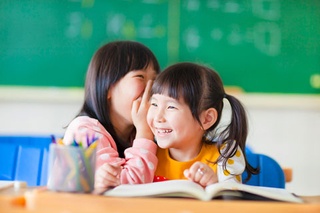 Two school girls are sitting at a desk with a textbook open in front of them. One girl is whispering in the other's ear and both are smiling and giggling.